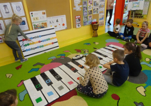 Dziewczynka wskazuje kolejność granych dźwięków na tablicy muzycznej, troje dzieci odtwarzają dźwięki na instrumencie.